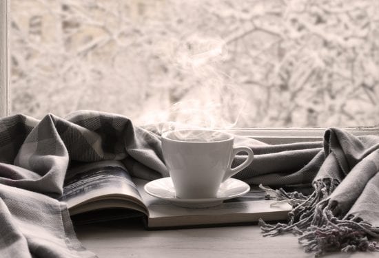 Cozy winter still life: cup of hot coffee and opened book with warm plaid on vintage windowsill against snow landscape from outside.